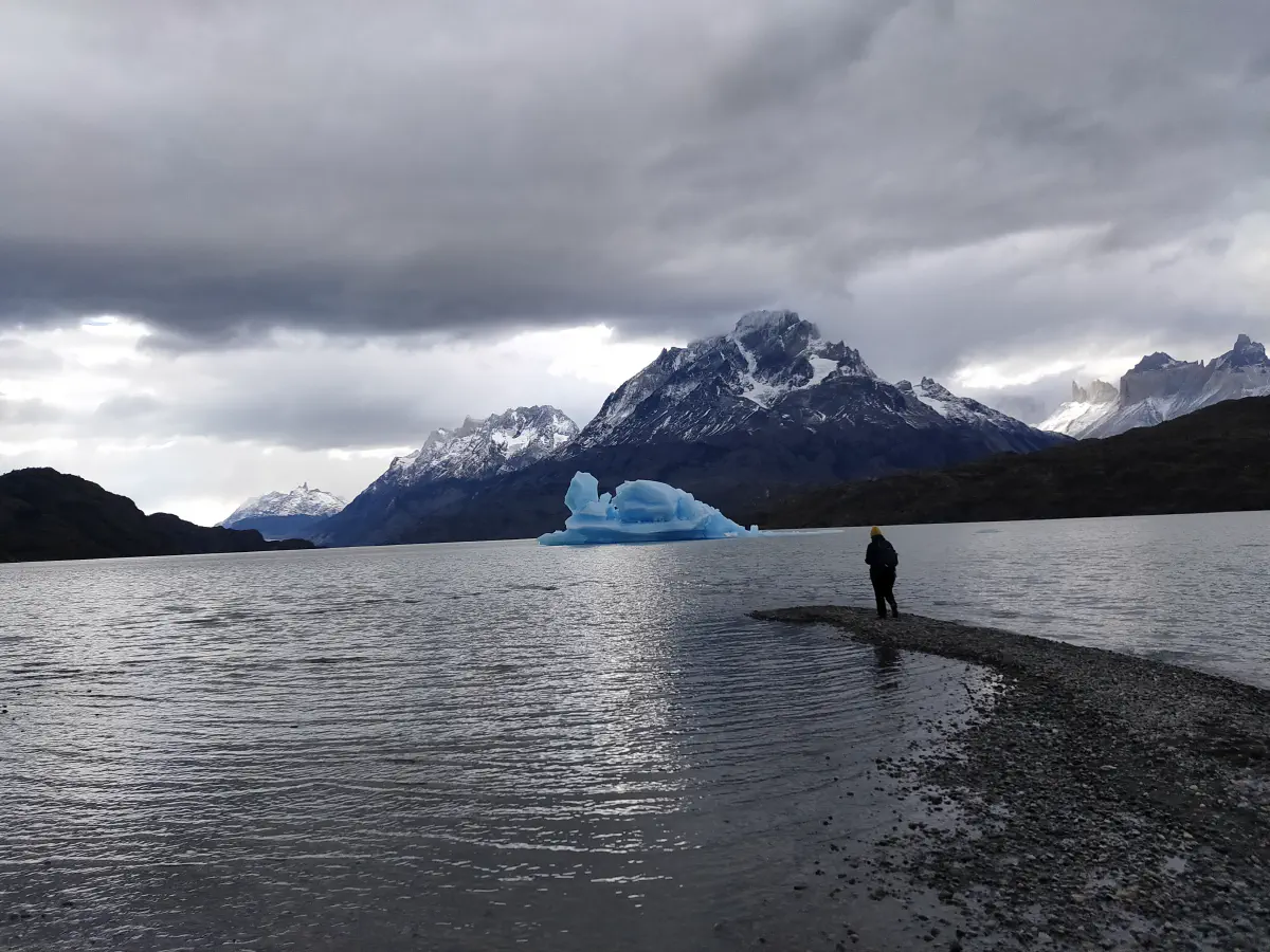 砂礫の岬に立つ人物と、ラゴ・グリスの湖面に浮かぶ青い氷山、背後に雪山